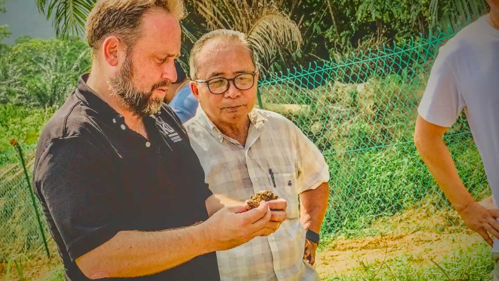 Man in foreground examining soil sample in his palms while another man in the background observes besides him.